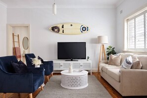 A living room with a TV, two chairs, a sofa, a round coffee table, and a surfboard decoration on the wall.