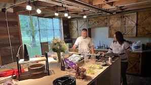 A rustic kitchen with wooden cabinets, a large island with food items, and a window overlooking greenery.