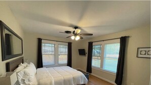 Une chambre à coucher avec un lit, un ventilateur de plafond et deux fenêtres équipées de stores.