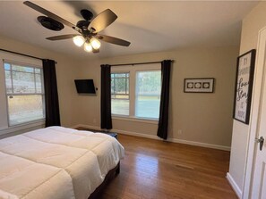 A bedroom with a bed, a ceiling fan, a TV, and windows with curtains.