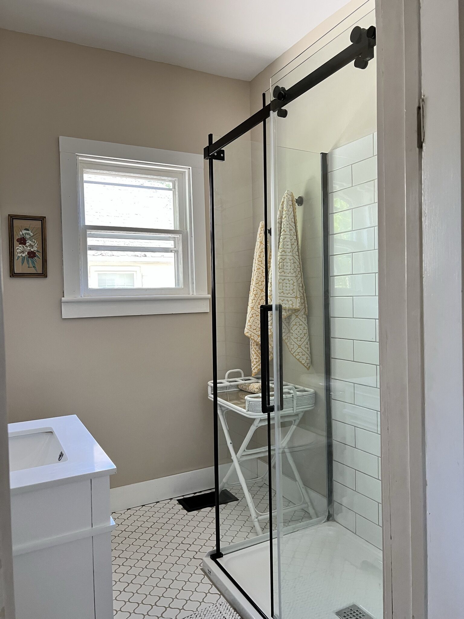 Une salle de bain avec une douche à parois de verre, un lavabo blanc, une fenêtre et un porte-serviettes.