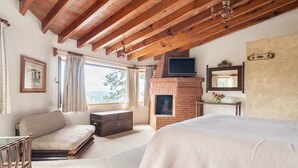 Quarto com cama, sofá, teto de madeira, lareira, televisão e janela com cortinas.