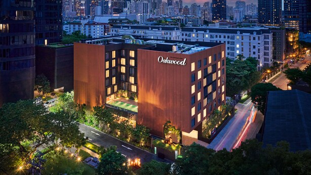 A modern building with the name "Oakwood" on its facade, surrounded by a well-maintained garden and a busy street. - Oakwood Studios Sukhumvit Bangkok (Bangkok)