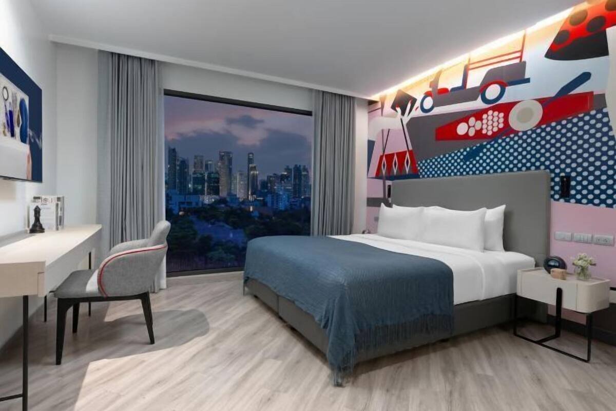 A modern hotel room with a large bed, a desk, and a chair, a city view through the window, and a wall with a colorful design.