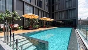A rooftop pool with clear water, surrounded by lounge chairs and umbrellas, and a modern building with large windows in the background. - Oakwood Studios Sukhumvit Bangkok (Bangkok)