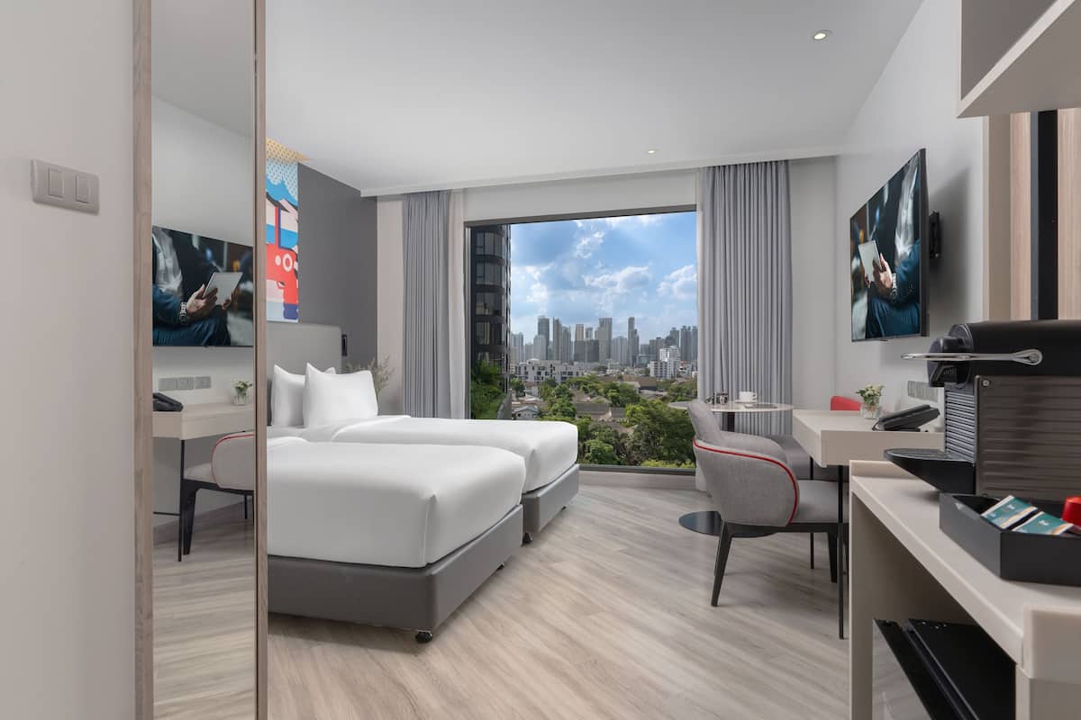 A modern hotel room with a city view, a desk with a laptop, a bed, and a seating area with a chair.