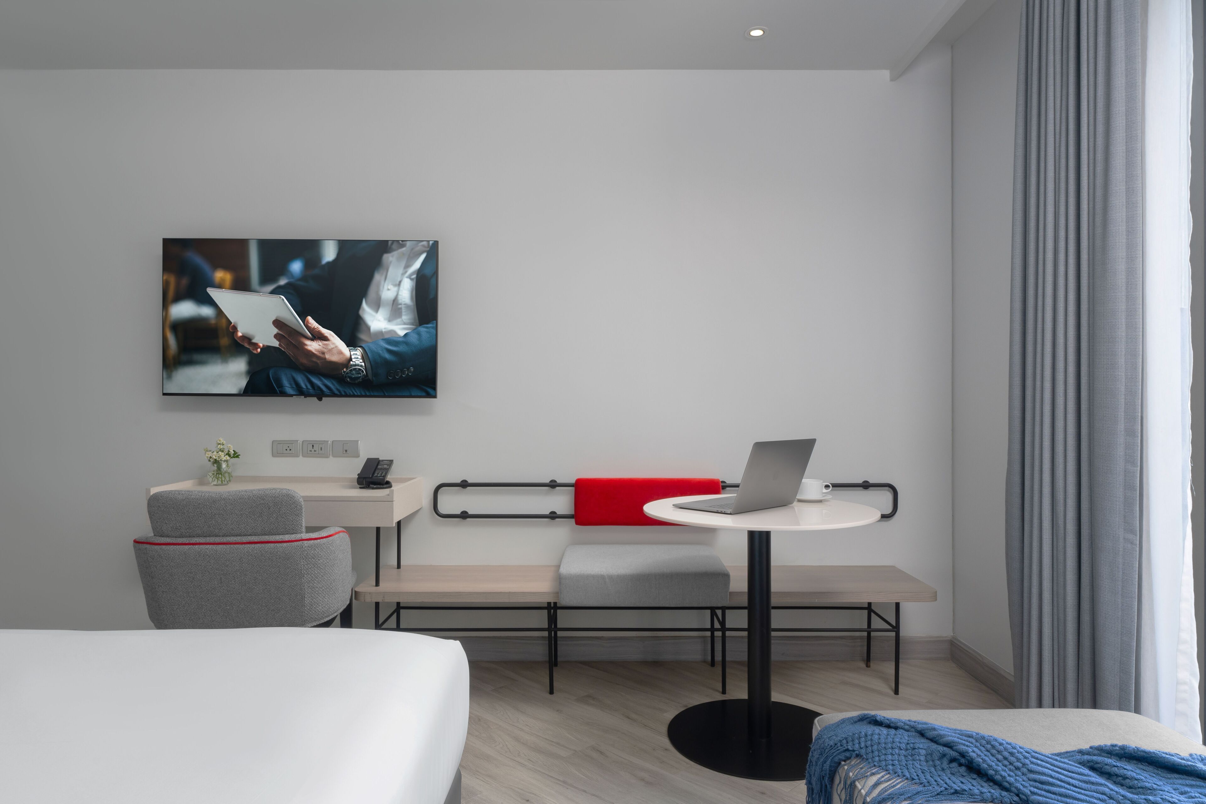 A modern hotel room with a bed, a desk with a laptop, a chair, a bench, and a wall-mounted TV.