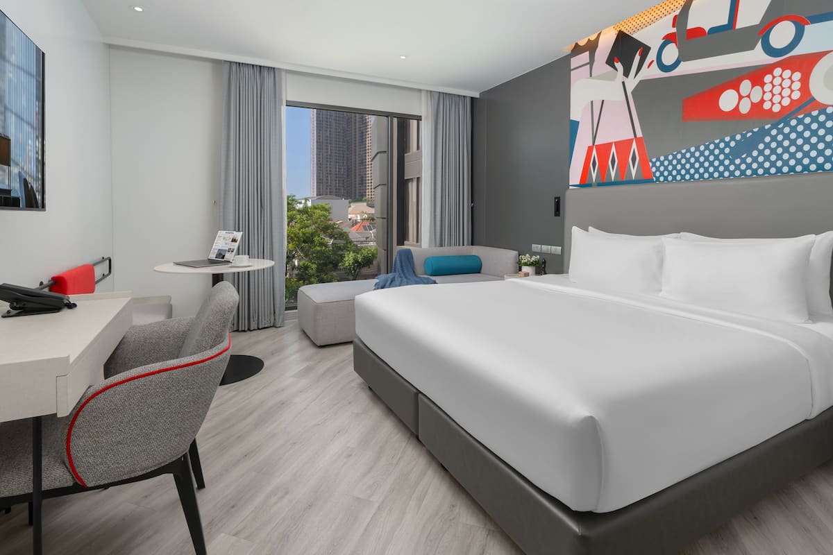A modern hotel room with a large bed, a desk, a chair, a small table, and a view of the city.