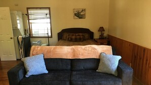 A room with a dark blue sofa, two matching pillows, a wooden coffee table, a bed with a plaid blanket, a deer painting on the wall, a lamp, and a window with blinds.