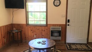 A room with a round table, a small side table, a heater, a clock, a television, and a door.