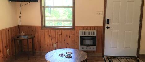 Una habitación con una mesa redonda, una mesita auxiliar, un calentador, un reloj, un televisor y una puerta.