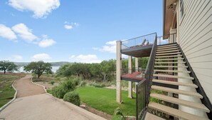 Una propiedad residencial con un sendero de hormigón que conduce a una zona ajardinada, un balcón con barandilla metálica y vistas a un lago y colinas.