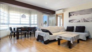 Un salón espacioso con piso de madera, un juego de sofás, una mesa de comedor con sillas y ventanas grandes con cortinas translúcidas.