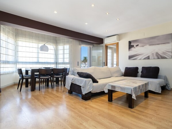 Un salón espacioso con piso de madera, un juego de sofás, una mesa de comedor con sillas y ventanas grandes con cortinas translúcidas.