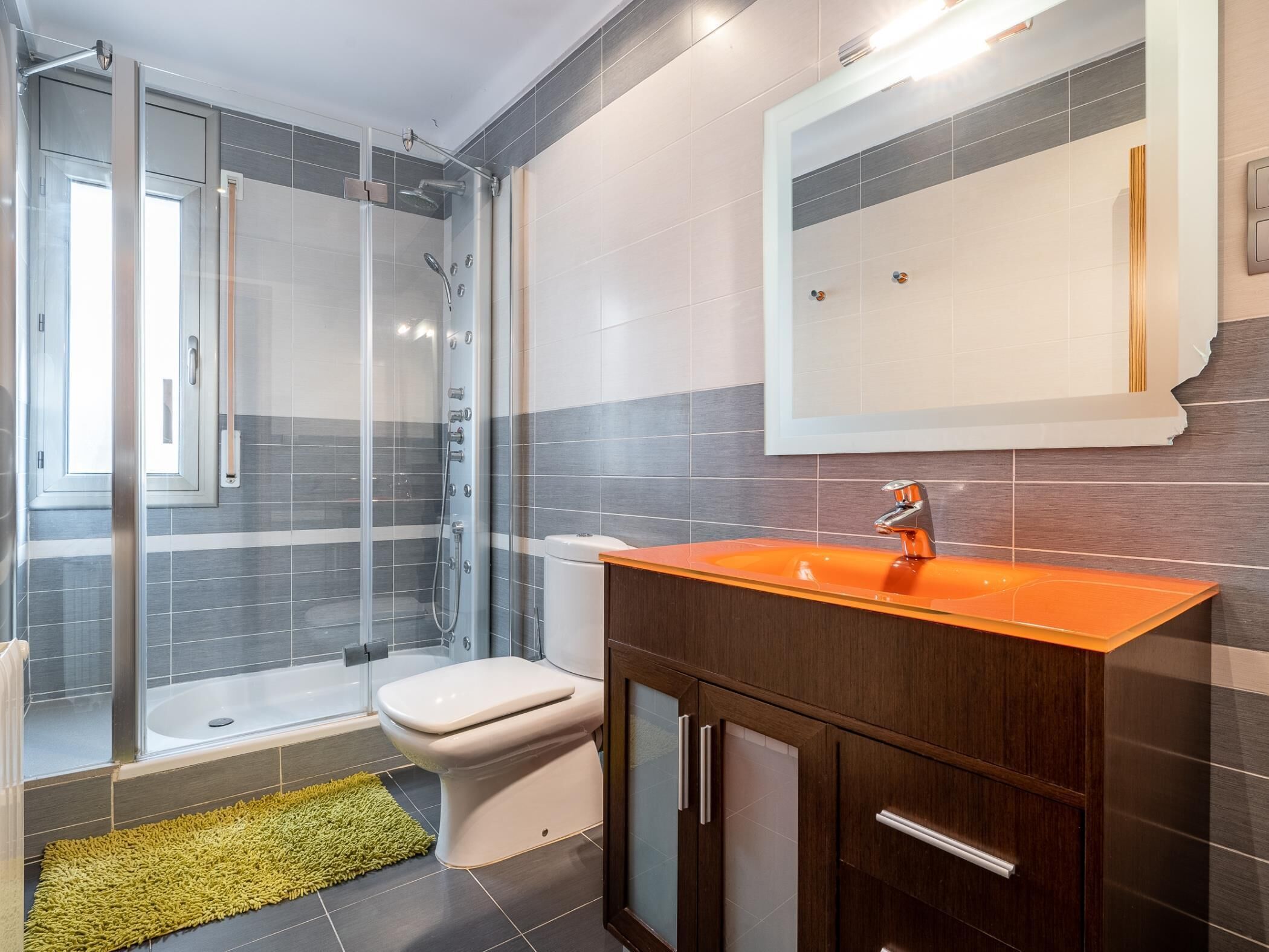 Un baño moderno con ducha envuelta en cristal, inodoro blanco y lavamanos naranja con muebles de madera.