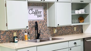 Een keuken met een betegelde spatwand, een bord met daarop 'Koffiebar' en diverse keukenapparaten.