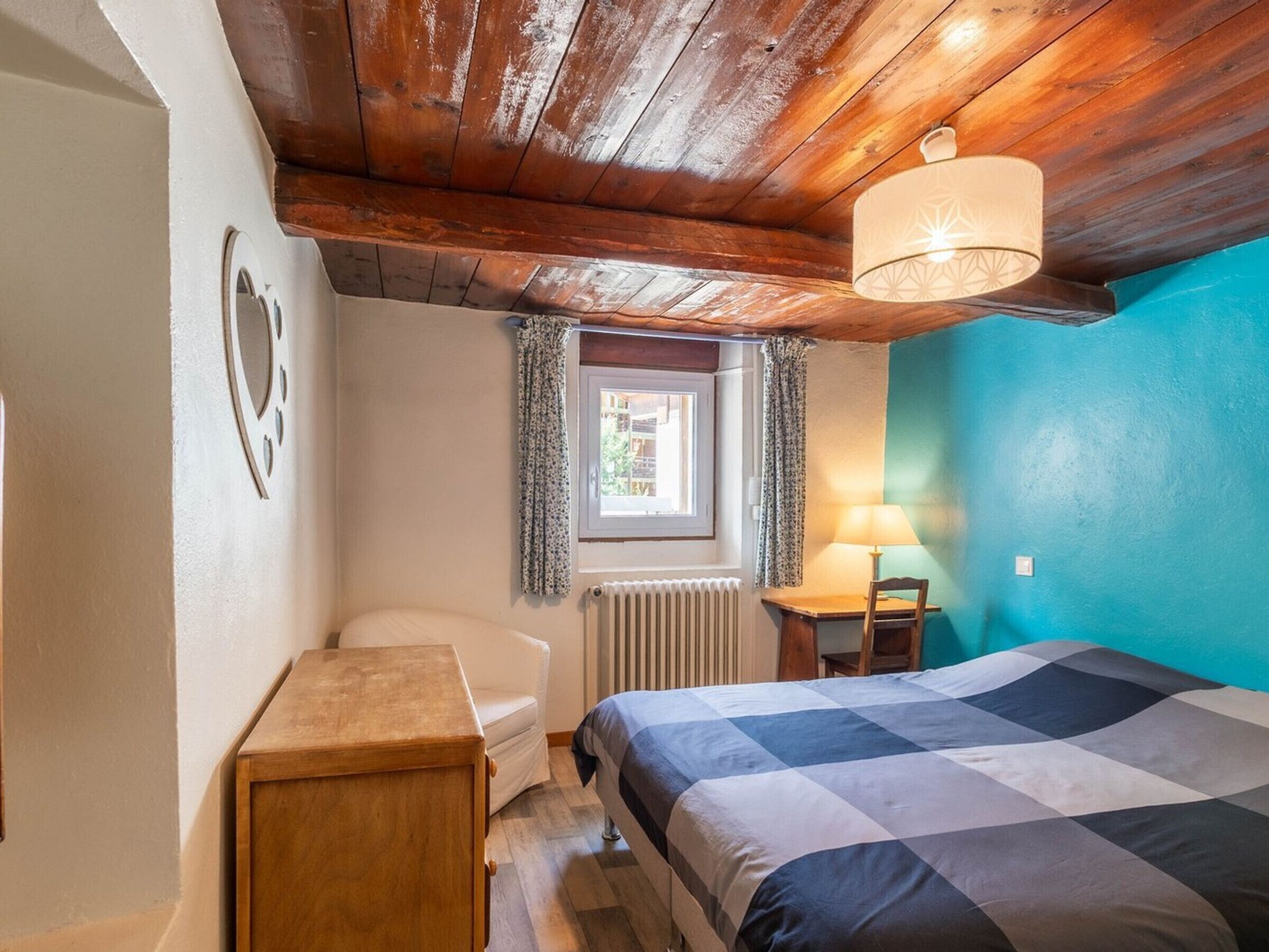 Un dormitorio con techo de madera, una cama con edredón a cuadros, una mesita de noche con lámpara, una ventana con cortinas y un radiador.