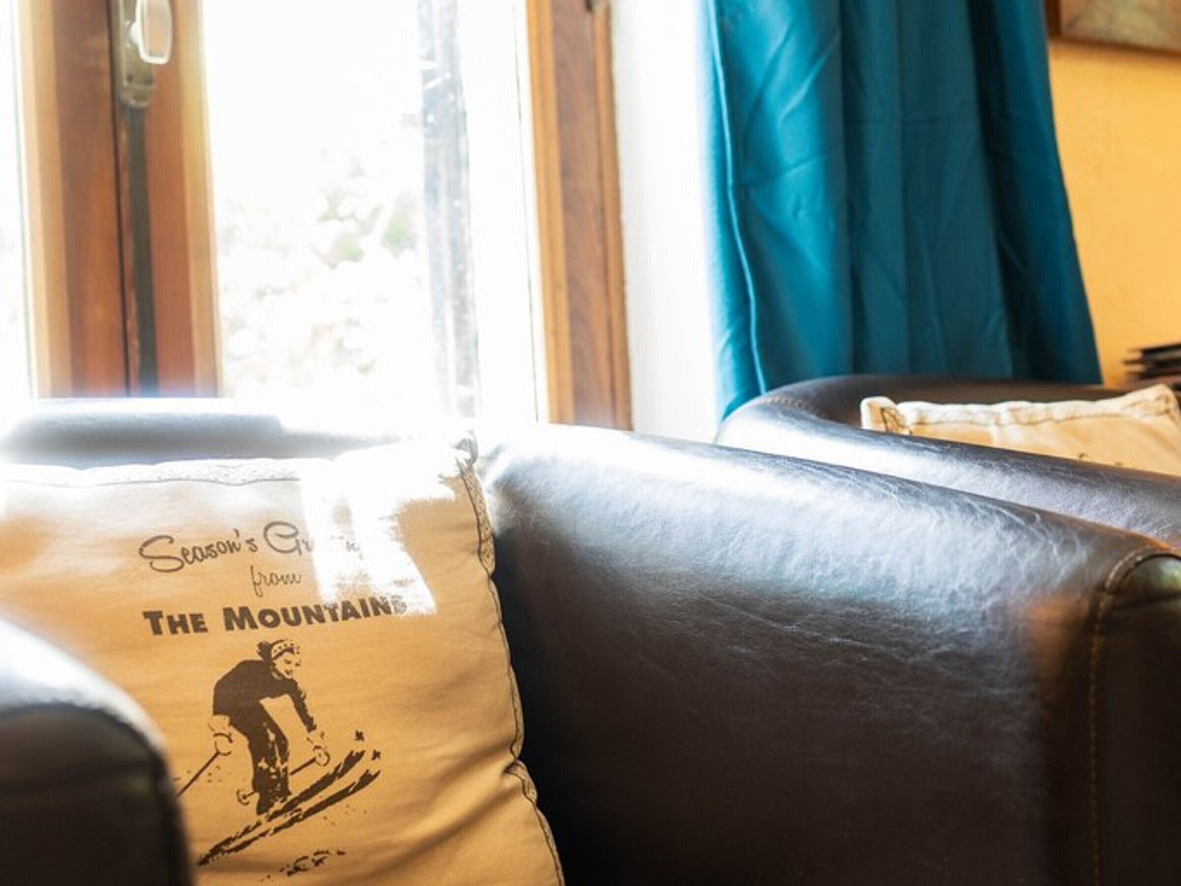 Ein gemütliches Zimmer mit Ledersofa, einem Fenster mit Vorhängen und einem Kopfkissen mit einem Ski-Motiv.