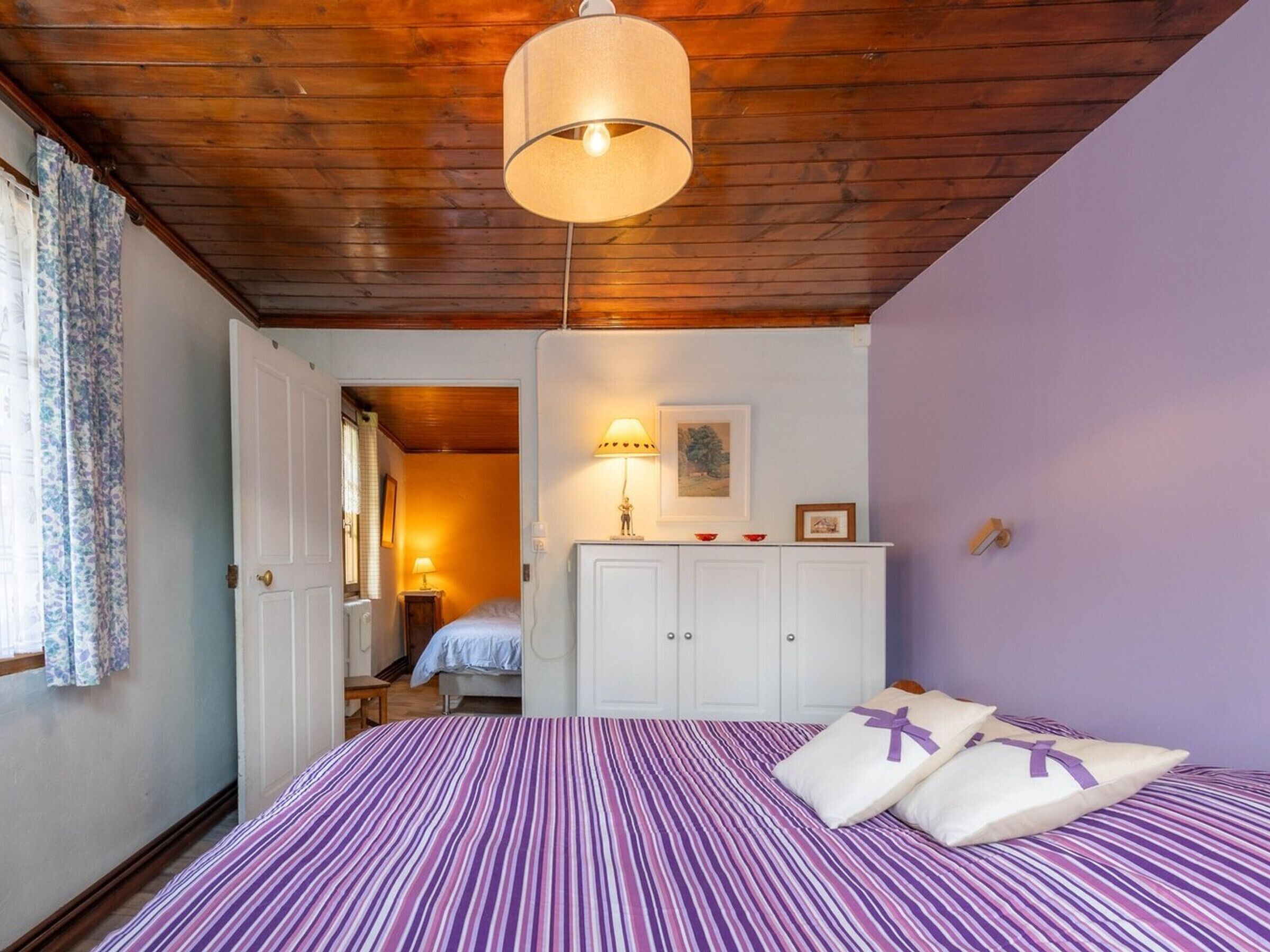 Un dormitorio con techo de madera, una cama con colcha a rayas, un tocador blanco y una lámpara colgante.