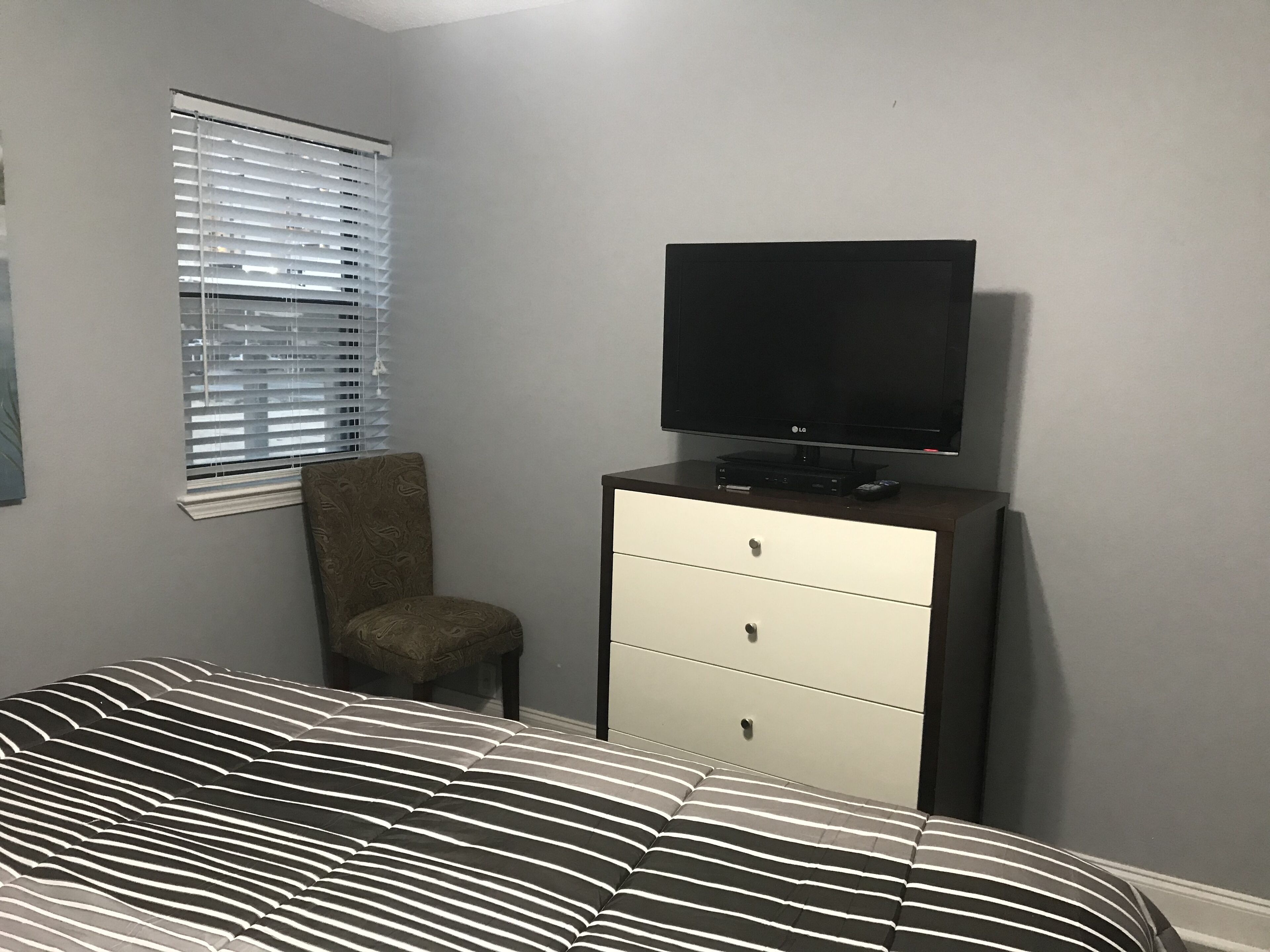 Een slaapkamer met een bed, een stoel, een dressoir en een televisie op een standaard.