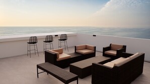 A rooftop terrace with wicker furniture, bar stools, and a view of the sea.