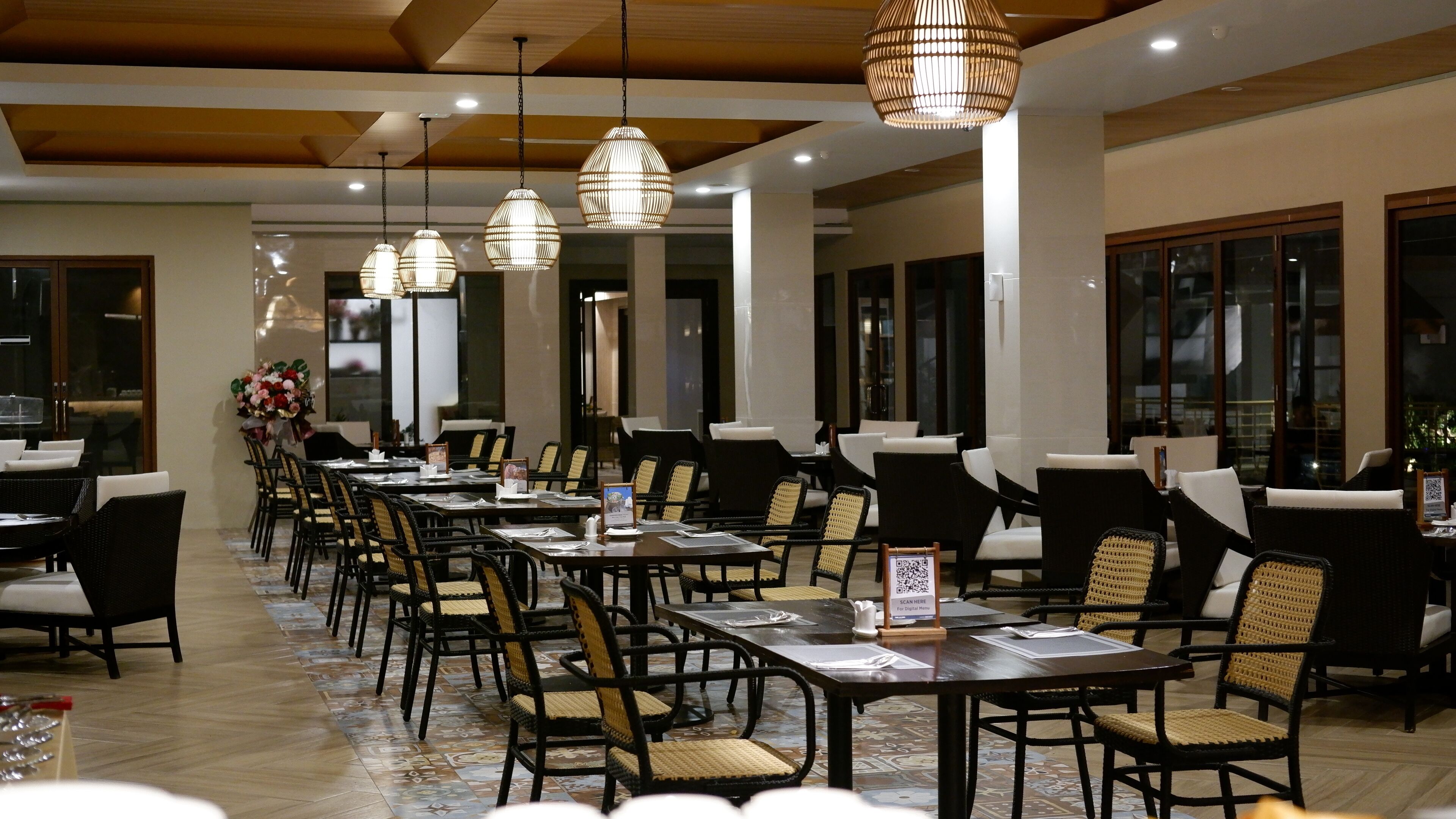 Ein Restaurant mit langen Tischen und Stühlen, hängenden Pendelleuchten und großen Fenstern.