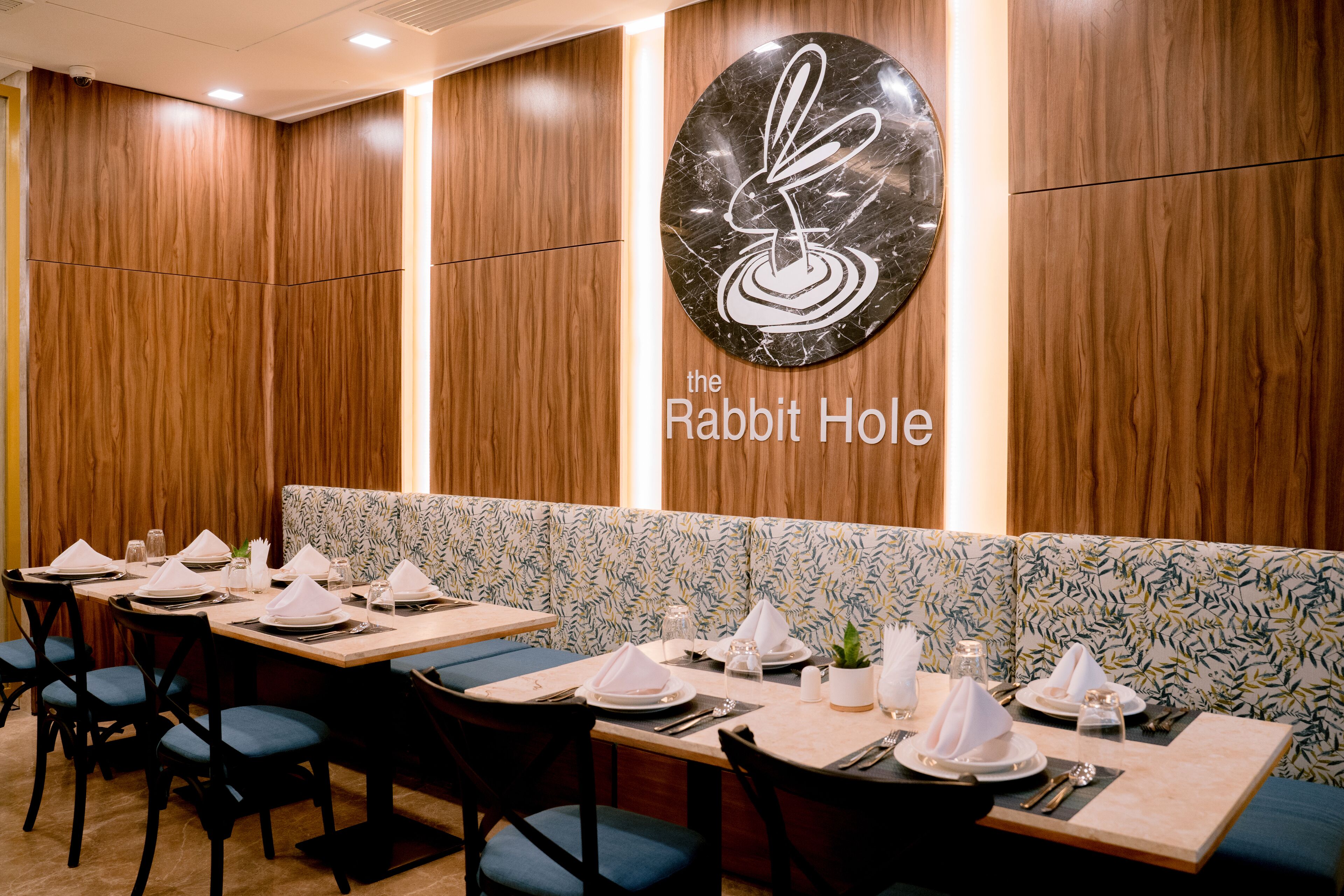 A restaurant interior with wooden paneling, a logo on the wall, and neatly set tables with blue chairs.