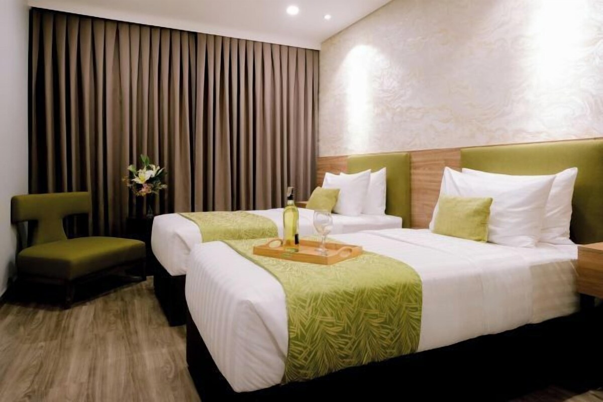 Habitación de hotel con dos camas, un sillón verde, una mesita de noche de madera y una bandeja decorativa con una botella y un vaso.