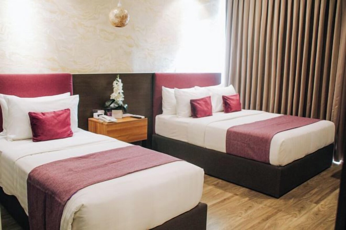 Habitación de hotel con dos camas, cada una con sábanas blancas y detalles rojos, una mesita de noche de madera y una planta decorativa.