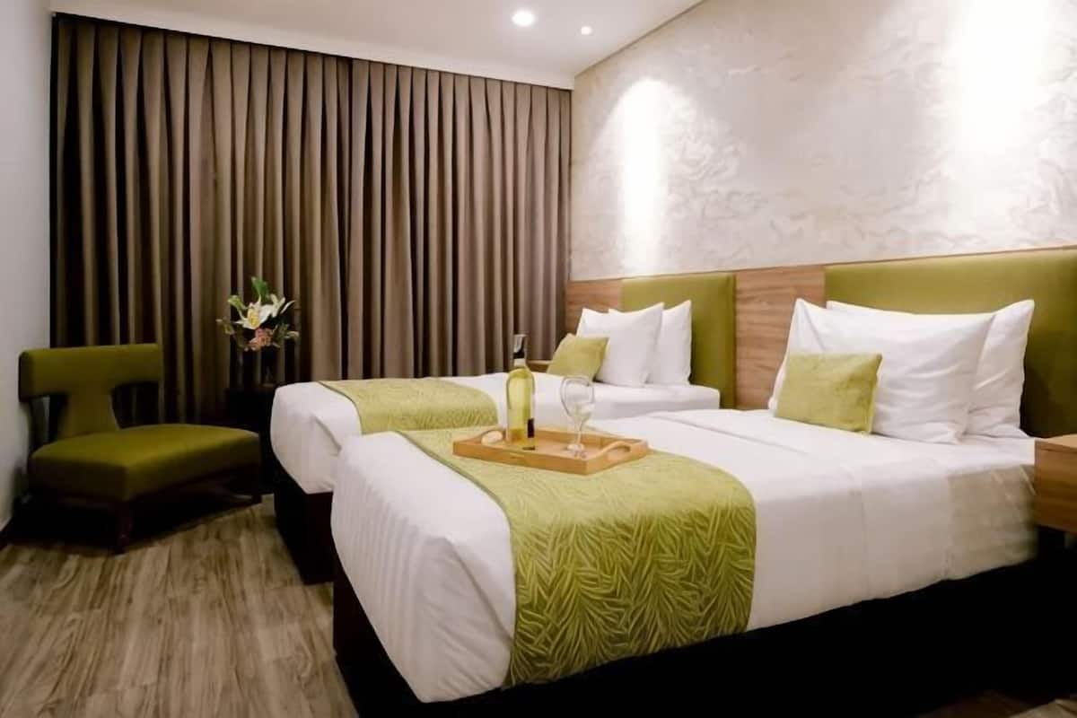 A hotel room with two beds, a green chair, a wooden nightstand, and a decorative vase.