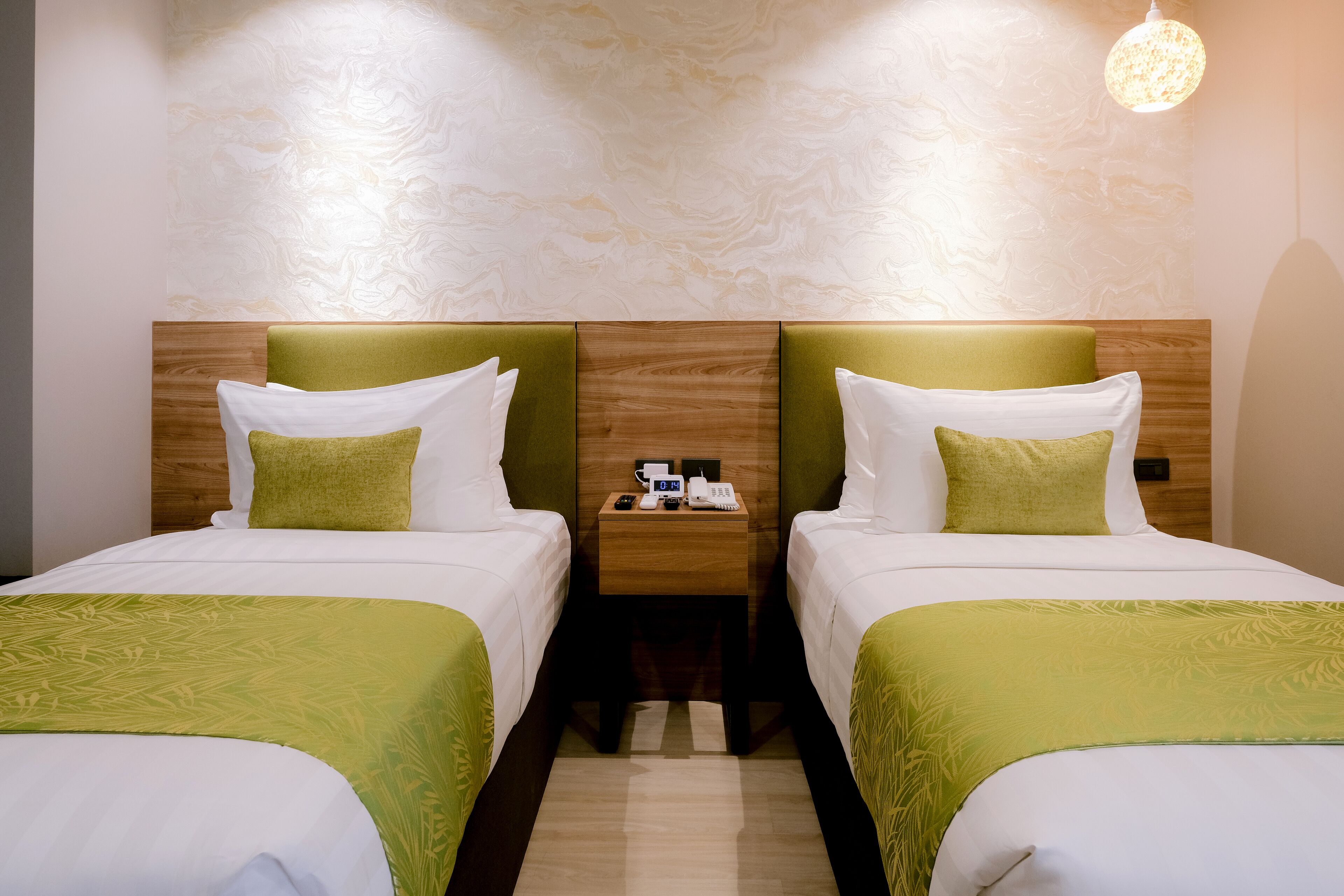 A hotel room with two beds, each with a green bedspread and white pillows. There is a wooden headboard, a nightstand with a phone and a lamp, and a marble-patterned wall.