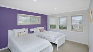 Quarto com duas camas, roupa de cama branca, paredes roxas e janelas grandes com persianas.