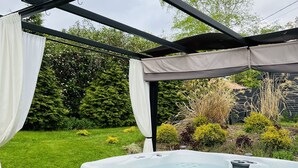 Un bain à remous installé sous une pergola agrémentée de rideaux blancs, entouré d’un jardin où poussent différentes plantes et d’une pelouse verdoyante.