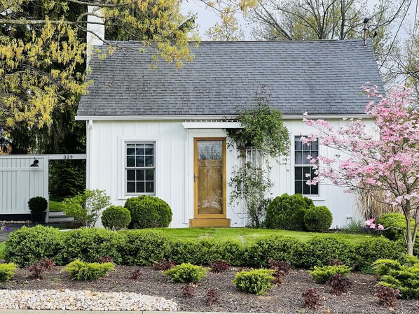 A white house with a dark roof, a wooden front door, and a well-maintained garden with hedges and a tree with pink flowers.