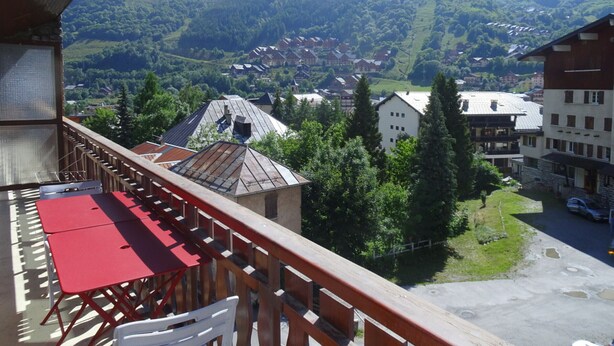 Ein Balkon mit rotem Tisch und weißem Stuhl, mit Blick auf ein Dorf mit Häusern und Bergen im Hintergrund.