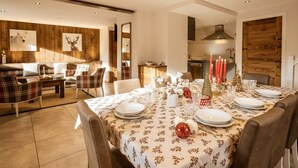 A dining area with a table set for six, surrounded by chairs. The room features wooden paneling on the walls and a modern kitchen in the background.