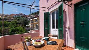 Un tavolo di legno con una ciotola di frutta e un libro su un balcone con una porta verde e finestre dotate di persiane.