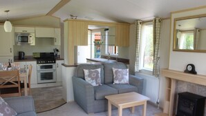 A compact living space with a kitchenette, a grey sofa with floral cushions, a wooden coffee table, and a fireplace. - Mountain View - luxury cabin with spectacular alpine and loch views (Taynuilt)