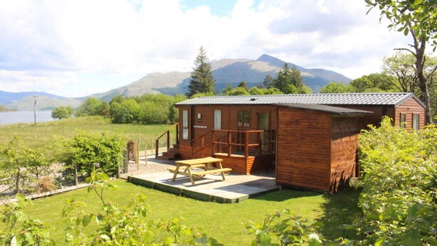 A wooden cabin with a patio and a view of a lake and mountains. - Mountain View - luxury cabin with spectacular alpine and loch views (Taynuilt)