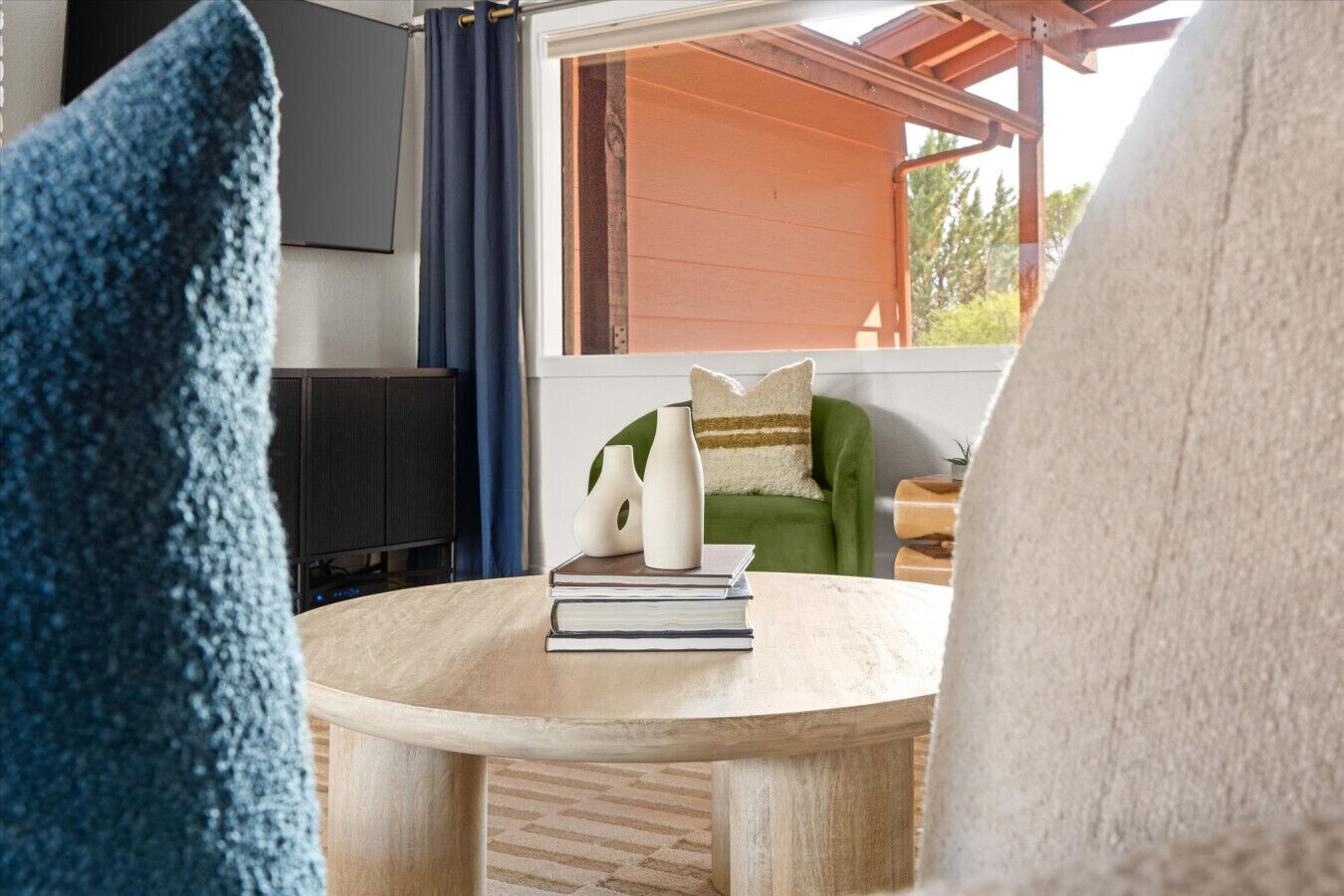 A round wooden table with a stack of books and two white vases. A green armchair with a striped pillow is visible in the background.