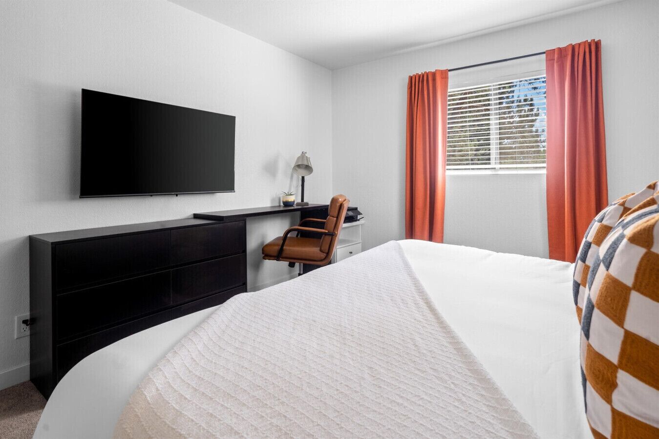 A hotel room with a bed, a desk with a chair, a television, and a window with curtains.