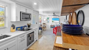 A modern kitchen with white cabinetry, a microwave, and a stove. There is a dining area with a table and chairs, and a view of the beach through the window. - Blue Egret: Pool, Enclosed Lanai, Close to the Beach by Anna Maria Life Vacation Rentals (Holmes Beach)