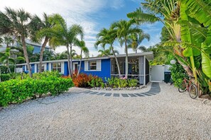 Une maison bleue avec un porche couvert, entourée de plantes tropicales et bordée d’une allée en gravier.