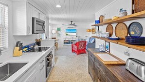 A modern kitchen with white cabinetry, a central island, and a dining area with a red chair. - Blue Egret: Pool, Enclosed Lanai, Close to the Beach by Anna Maria Life Vacation Rentals (Holmes Beach)