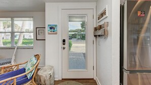 A compact kitchen with a refrigerator, a dining area with a chair and table, and a door leading to a covered outdoor area. - Blue Pelican: Sparkling Pool, Patio Dining, Close to the Beach by Anna Maria Life Vacation Rentals (Holmes Beach)