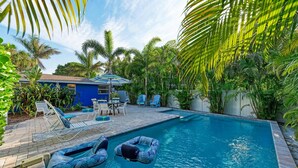 A swimming pool with lounge chairs and inflatable rings, surrounded by palm trees and a blue building. - Blue Pelican: Sparkling Pool, Patio Dining, Close to the Beach by Anna Maria Life Vacation Rentals (Holmes Beach)