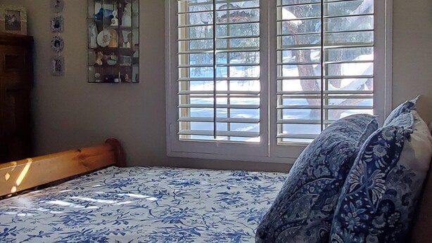 Une chambre à coucher avec un lit, une fenêtre avec des stores, une tête de lit en bois et une étagère murale agrémentée d’objets décoratifs.