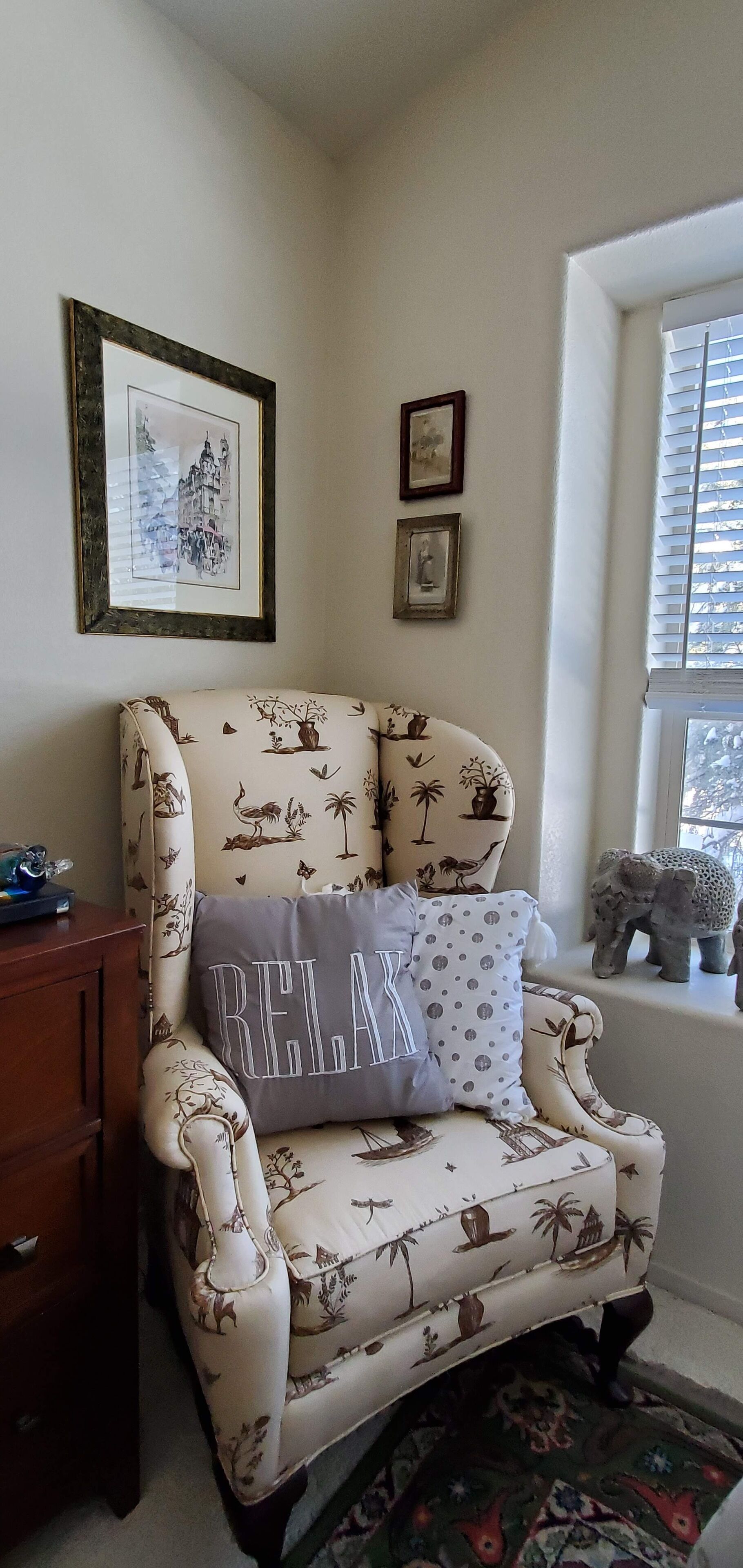 Une pièce d’angle avec un fauteuil à motifs, un tableau encadré au mur et une fenêtre avec des stores.