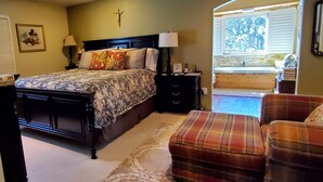 Une chambre à coucher avec un lit, des tables de chevet, une croix accrochée au mur, une fenêtre donnant sur un paysage et une chaise.