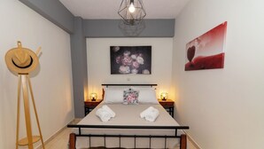Cama bem arrumada com lençóis brancos e duas toalhas enroladas, um cavalete de madeira e duas luminárias fixadas na parede, uma de cada lado.